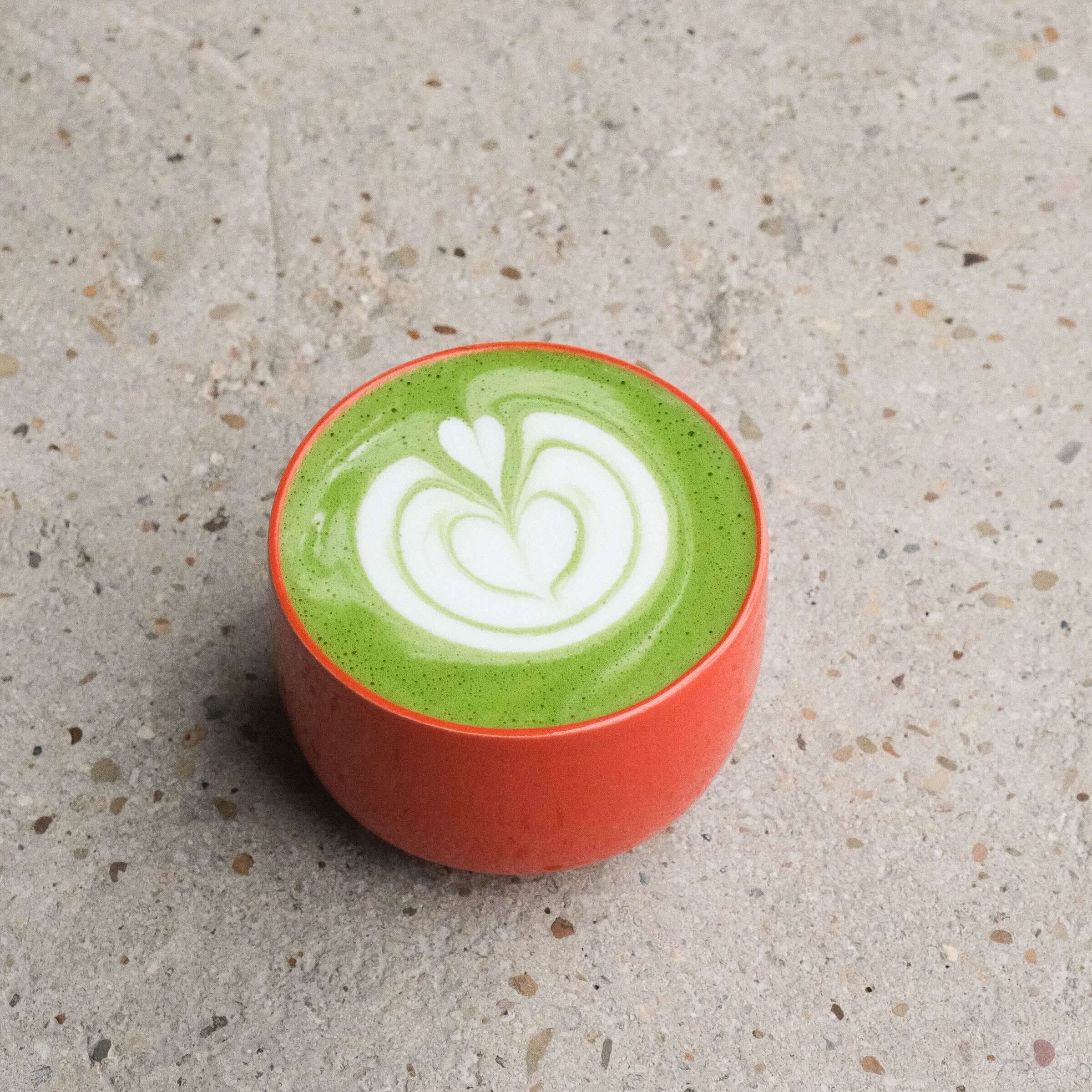 Vibrant green matcha latte with intricate white latte art in a glossy red cup on a textured surface.