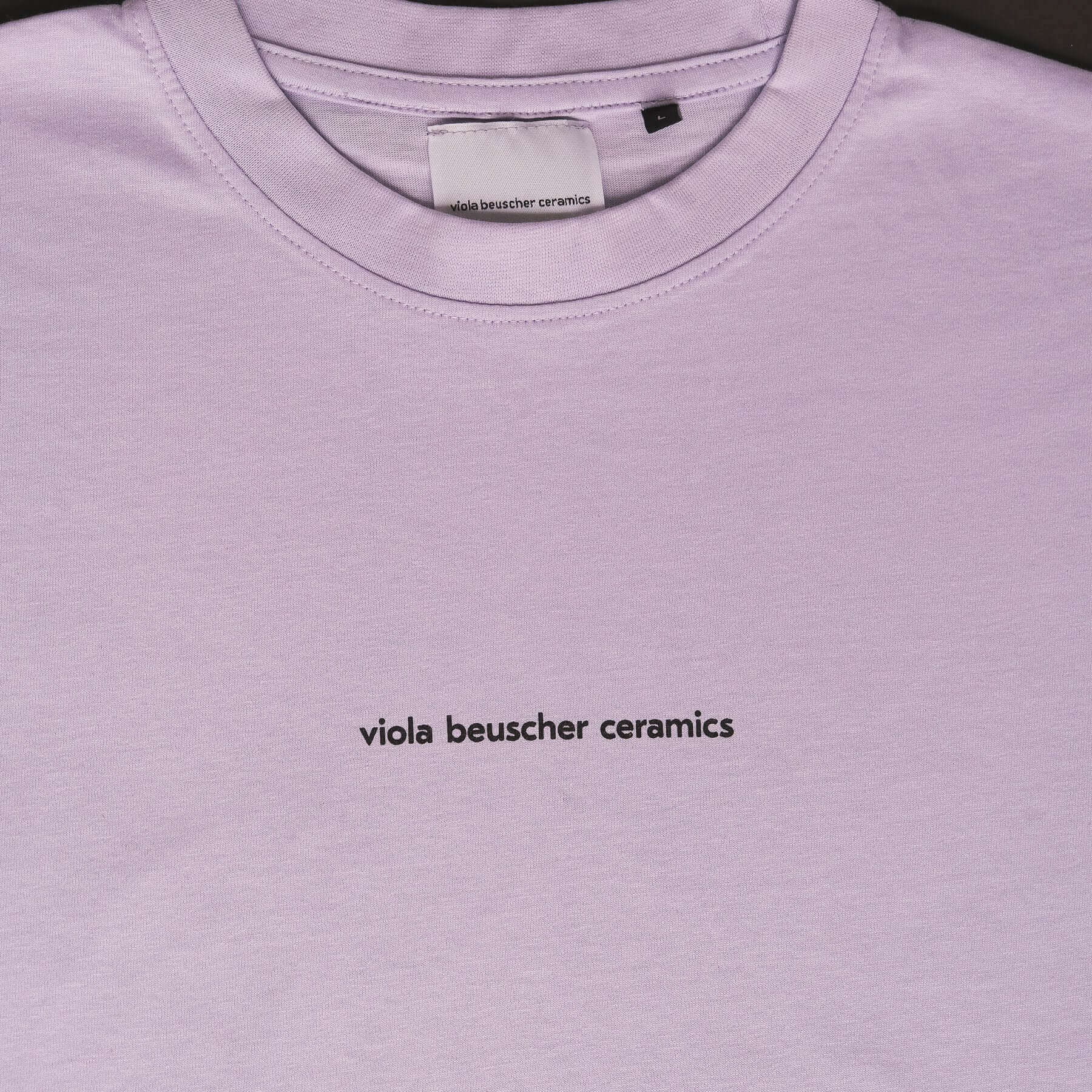 Close-up of lilac T-shirt featuring black text 'viola beuscher ceramics' on the front