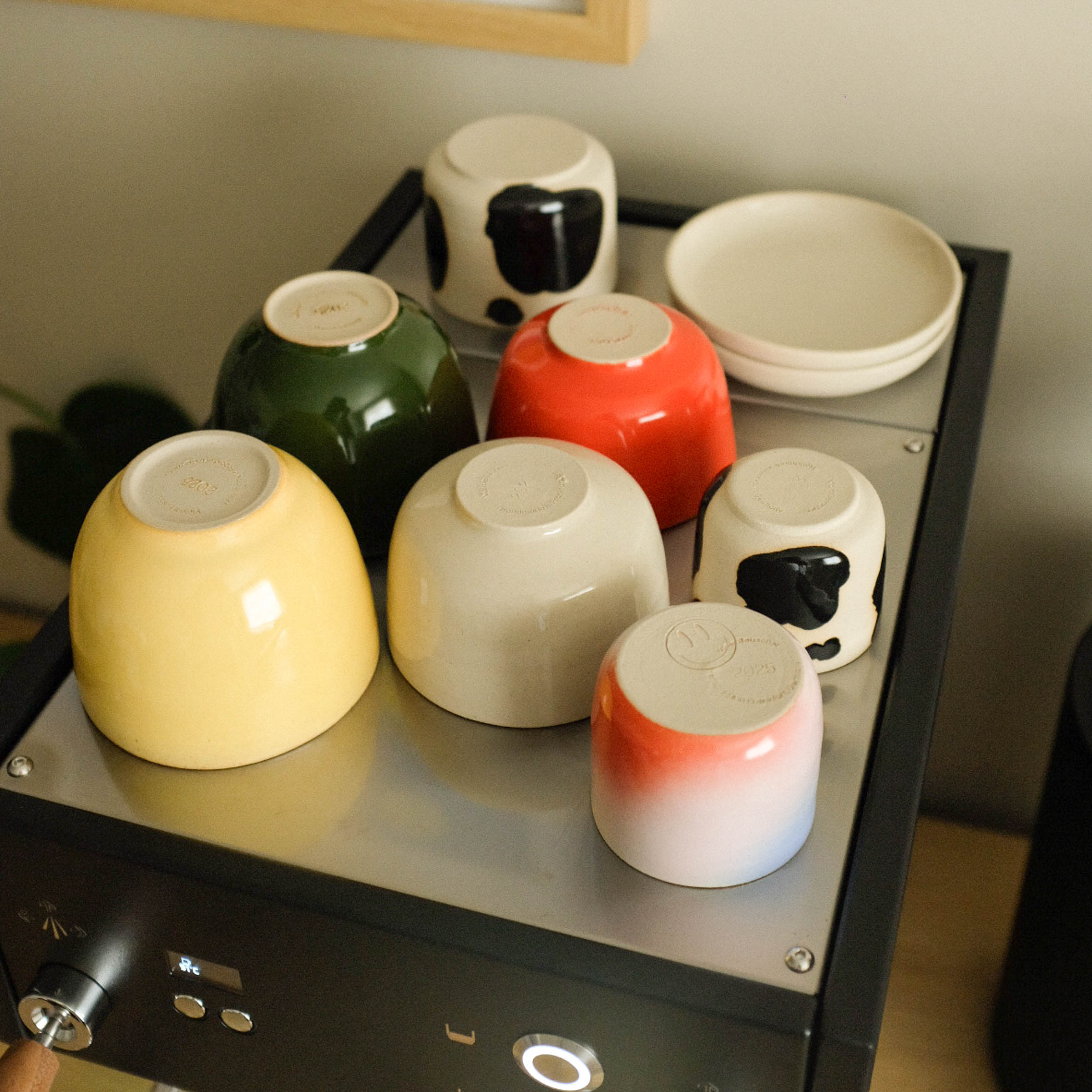Assorted colorful stoneware cups and bowls arranged on a coffee machine, showcasing modern ceramic craftsmanship.