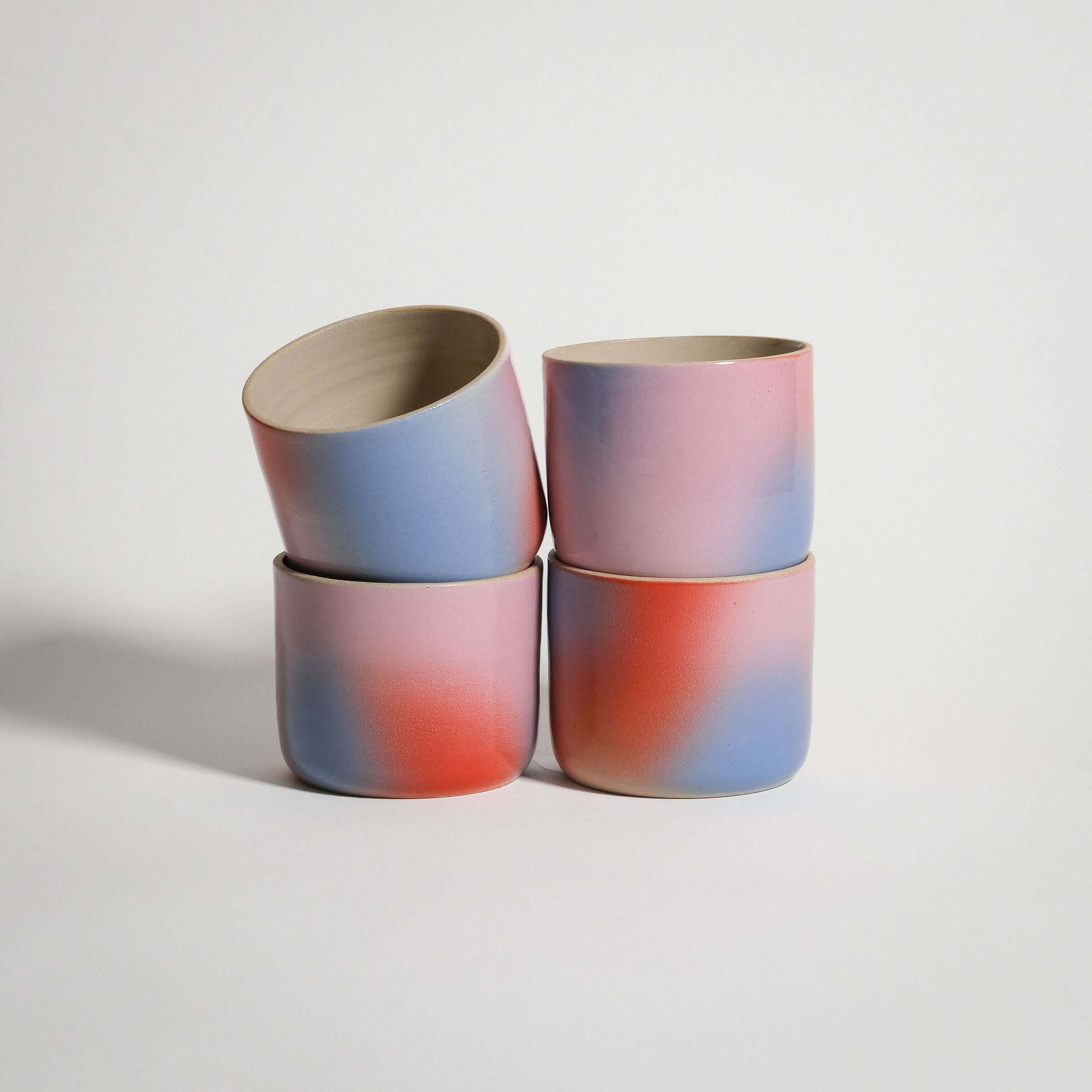 Handcrafted grey stoneware coffee cups with a gradient glaze in red, blue, and lilac, stacked four together.