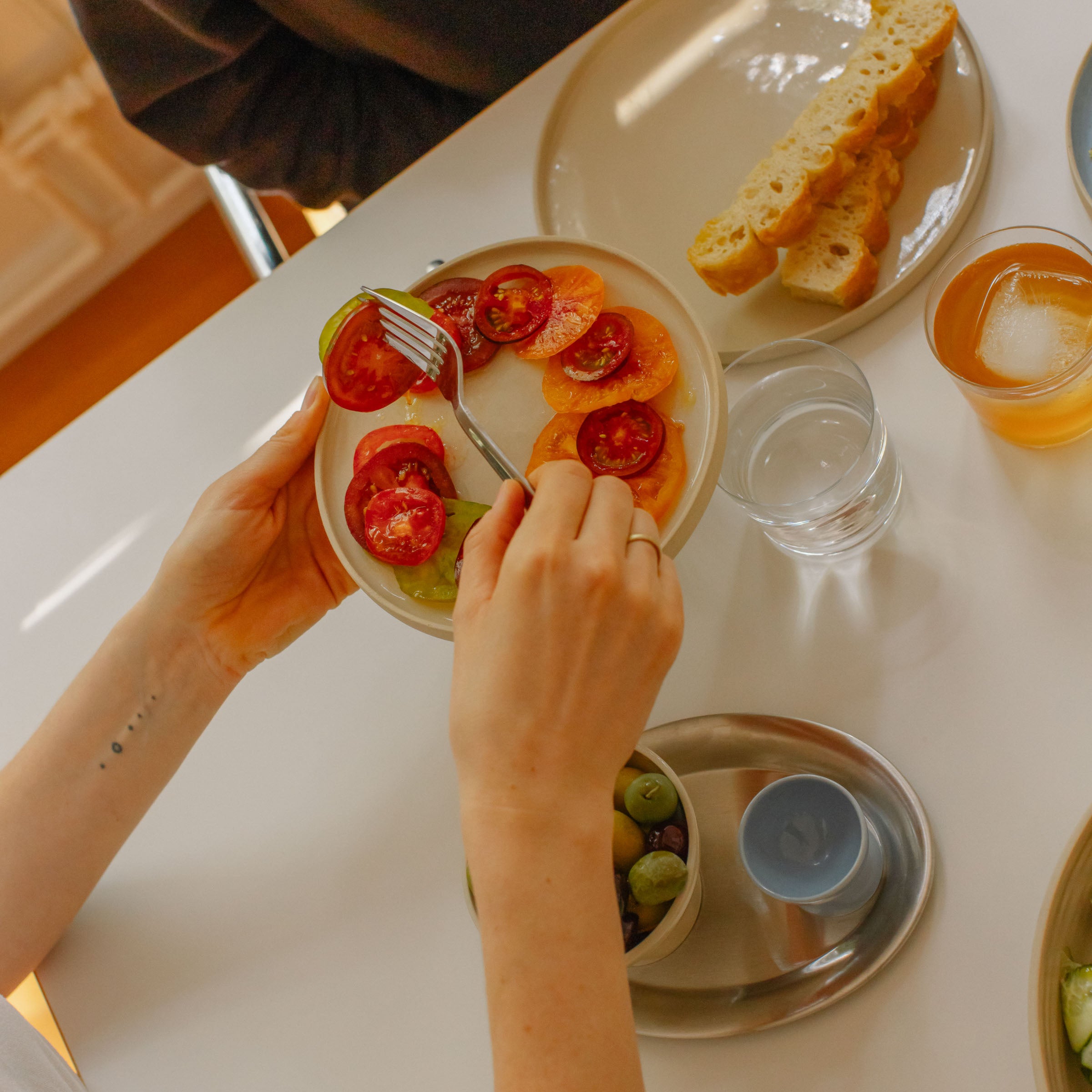 Hand holding a creme breakfast plate with sliced tomatoes, next to a plate of bread and drinks on a table.