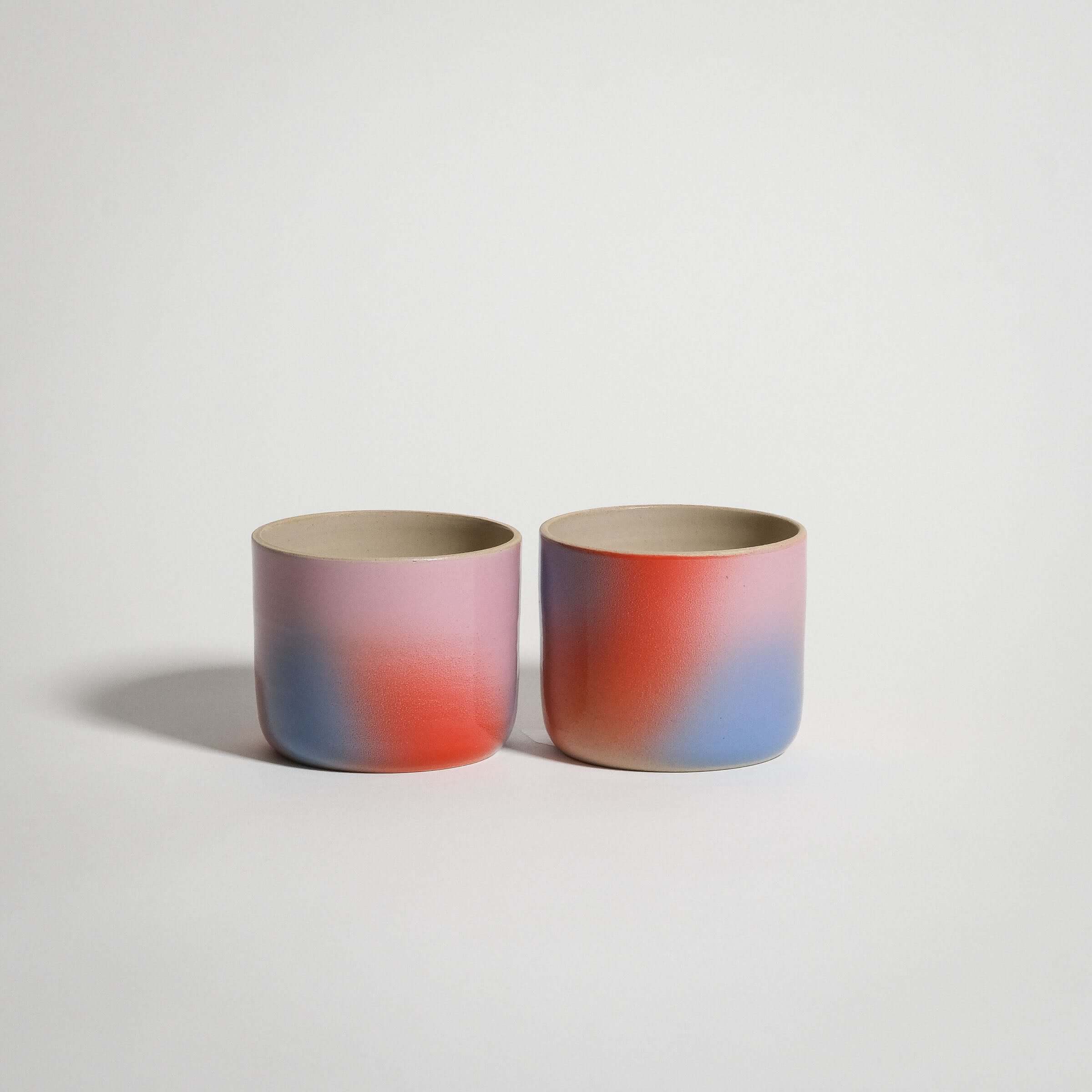 Handcrafted grey stoneware coffee cups with a glossy glaze and spray-painted gradient in red, blue, and lilac.