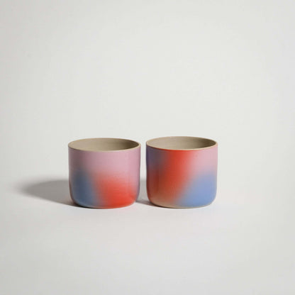 Handcrafted grey stoneware coffee cups with a glossy glaze and spray-painted gradient in red, blue, and lilac.