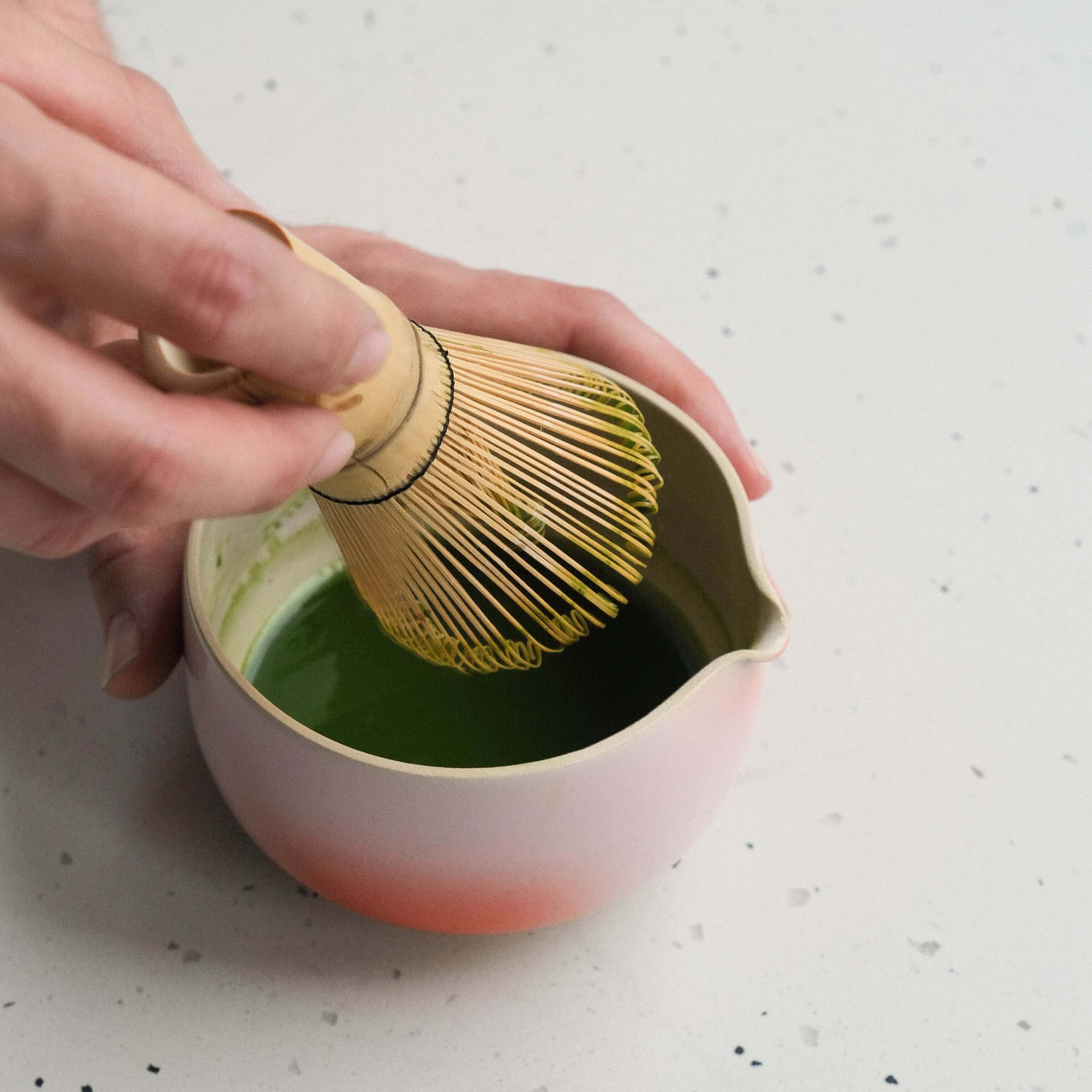 Hand using a bamboo whisk to mix matcha in a handmade pink and red pouring bowl.