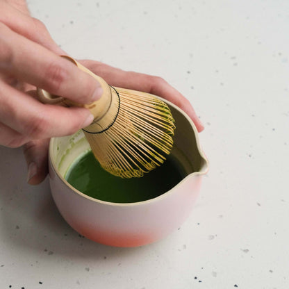 Hand using a bamboo whisk to mix matcha in a handmade pink and red pouring bowl.