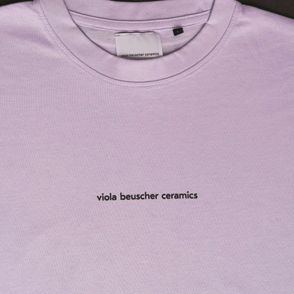 Close-up of lilac T-shirt featuring black text 'viola beuscher ceramics' on the front