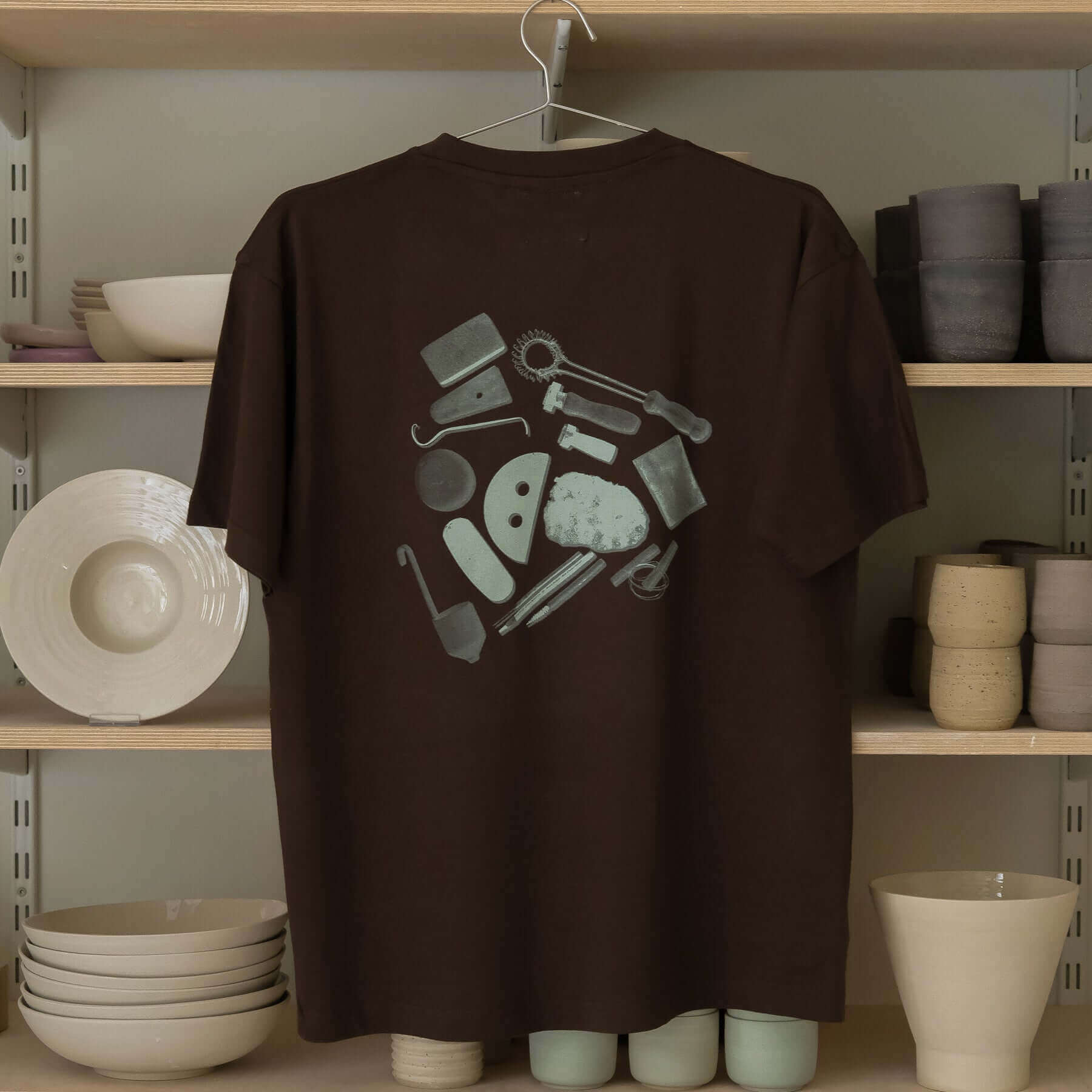 Back view of vbc Team Shirt in chocolate color featuring pottery tools print on a backdrop of ceramic items.