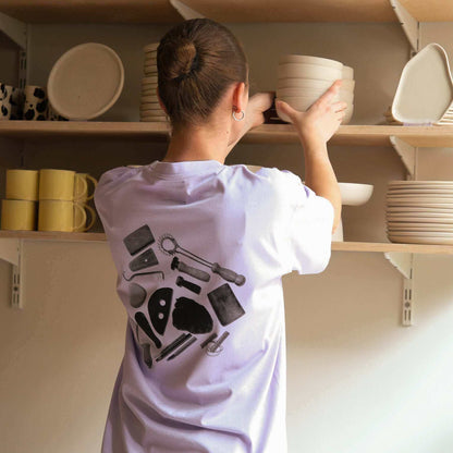 Person organizing pottery items on a shelf while wearing a lilac team shirt featuring tool designs on the back.