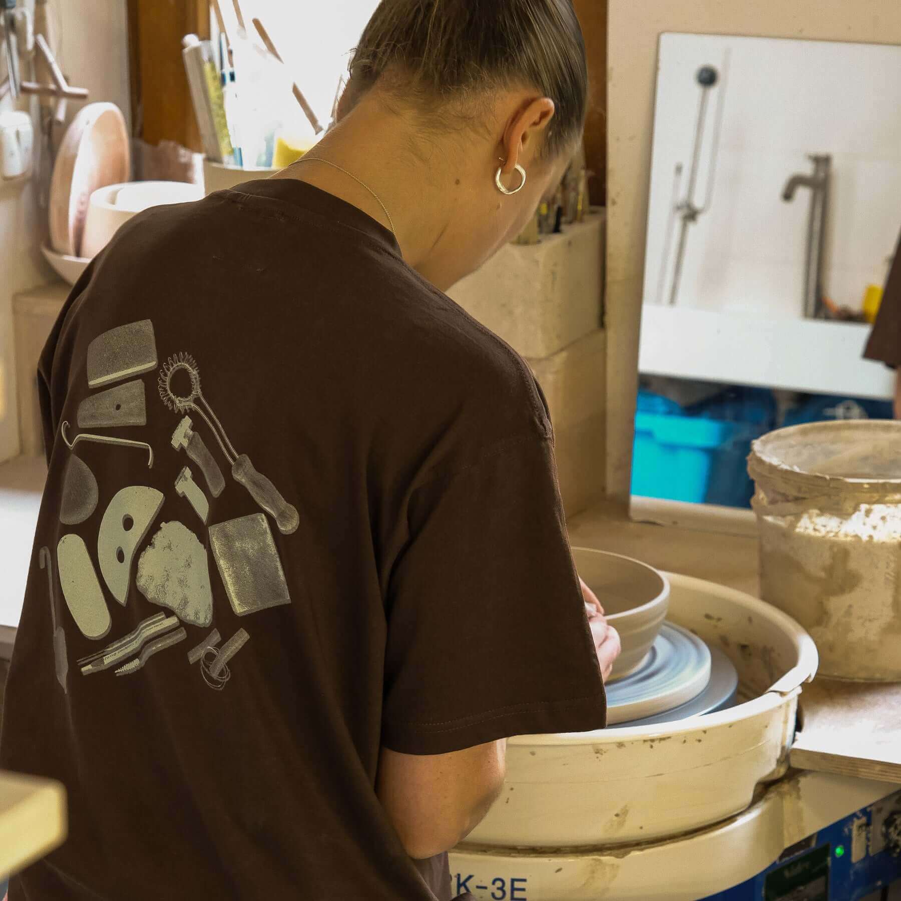Woman shaping pottery in a chocolate vbc team shirt with tool graphic on back, in a pottery studio.