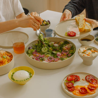 Handmade creme serving bowl filled with fresh salad, surrounded by plates of various dishes on a dining table.